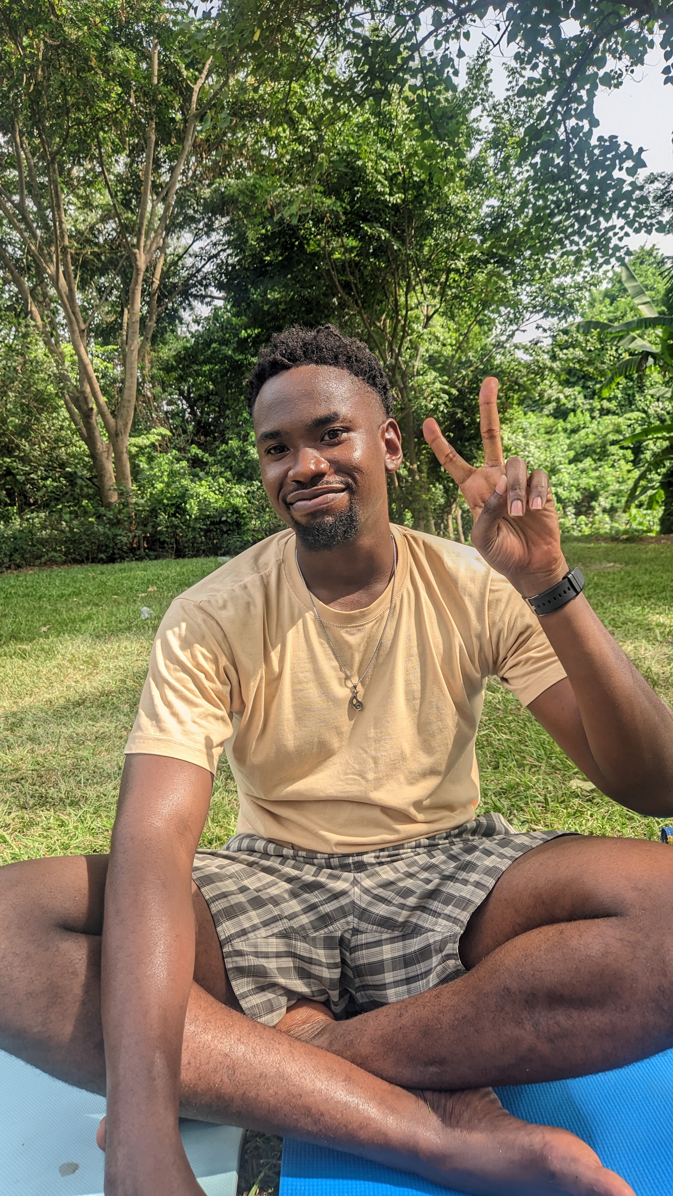 Mezuo smiling at the camera, sitting cross-legged over a green lawn. He is showing a victory sign with his left hand, over a background of trees and foliage.